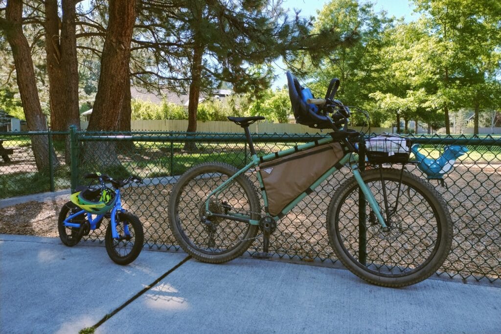 Surly Grappler with child seat attached to front of bike. Small child's bike next to it a playground.